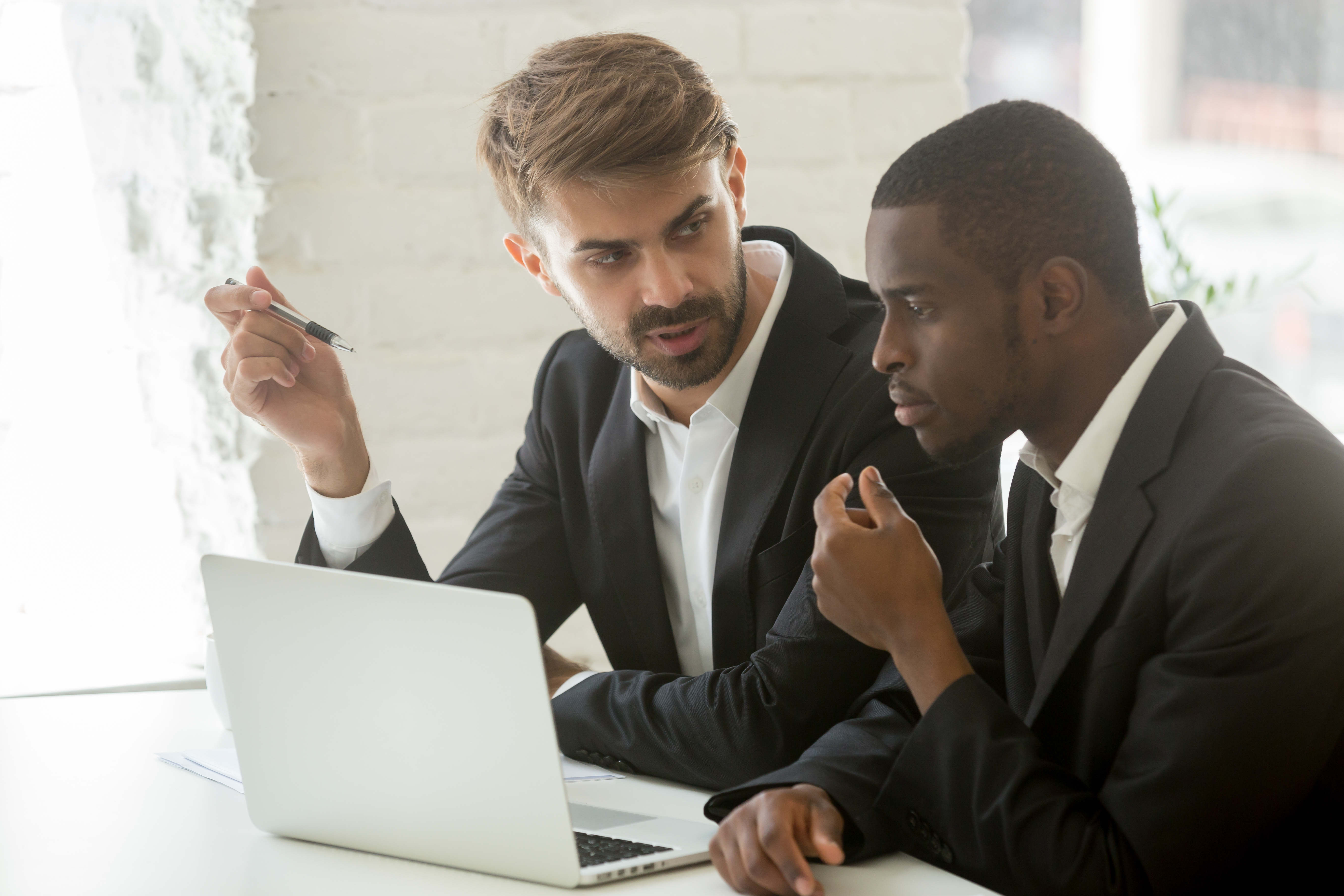 African and caucasian businessmen discuss online project idea with laptop, diverse executive partners in suits work together talking thinking about business improvement and stock trading investment
